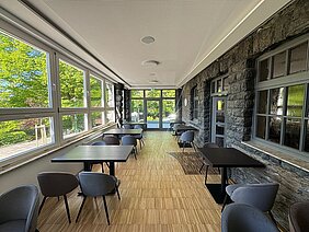 Heller Sitzbereich im sanierten Berggasthaus auf dem Pfaffenberg mit modernen Tischen, großen Fensterflächen und historischer Natursteinwand.