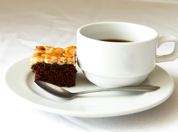 Weiße Kaffeetasse mit schwarzem Kaffee auf einem Unterteller, daneben ein Stück Schokoladenkuchen mit Nüssen und ein kleiner Löffel.