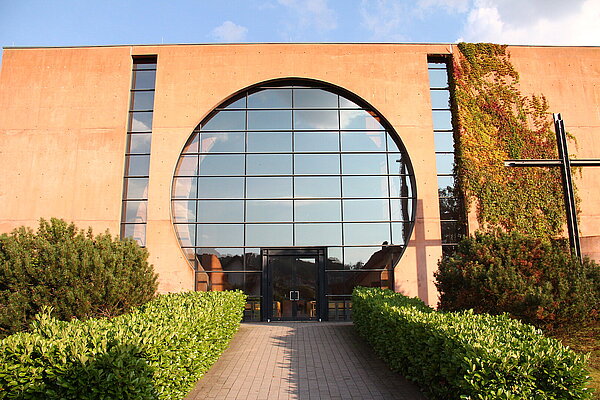 Außenansicht der katholischen Kirche St. Pius X. in Hohenstein-Ernstthal. Die moderne Fassade prägt ein großes, rundes Glasfenster über dem Eingang; seitlich wächst Efeu an der Wand. Ein von Hecken gesäumter Weg führt zum Gebäude. Die Kirche wurde von Peter Böhm entworfen und 1998 geweiht.