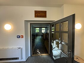 Eingangsbereich des sanierten Berggasthauses auf dem Pfaffenberg mit historischem Berggasthaus-Schild über der geöffneten Tür und Blick in die neu gestaltete Gaststube.
