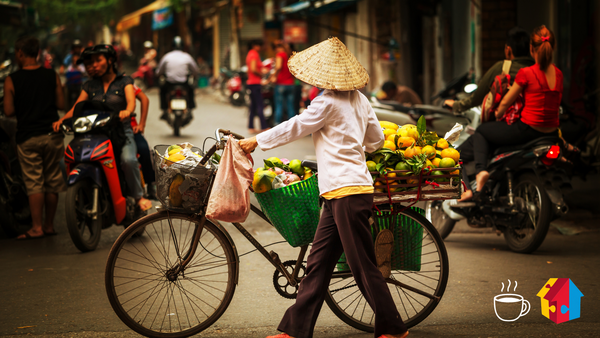 Eine vietnamesische Straßenverkäuferin mit traditionellem Kegelhut schiebt ein Fahrrad mit Körben voller frischer Früchte durch eine belebte Straße. Im Hintergrund sind Menschen auf Motorrollern und zu Fuß unterwegs. Unten rechts befinden sich ein Kaffeesymbol und das Logo des Mehrgenerationenhauses.