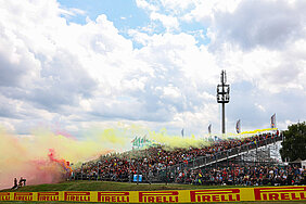 Dicht besetzte Zuschauertribüne am Sachsenring beim Motorrad Grand Prix mit farbigem Rauch in Gelb und Rot über den Fans – die Szene zeigt die lebendige Fankultur und besondere Atmosphäre der Rennstrecke in Hohenstein-Ernstthal.
