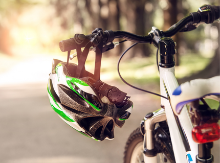 Nahaufnahme eines Fahrrads mit einem grün-weißen Fahrradhelm, der am Lenker hängt. Im Hintergrund ist unscharf ein sonniger Weg zwischen Bäumen zu sehen.